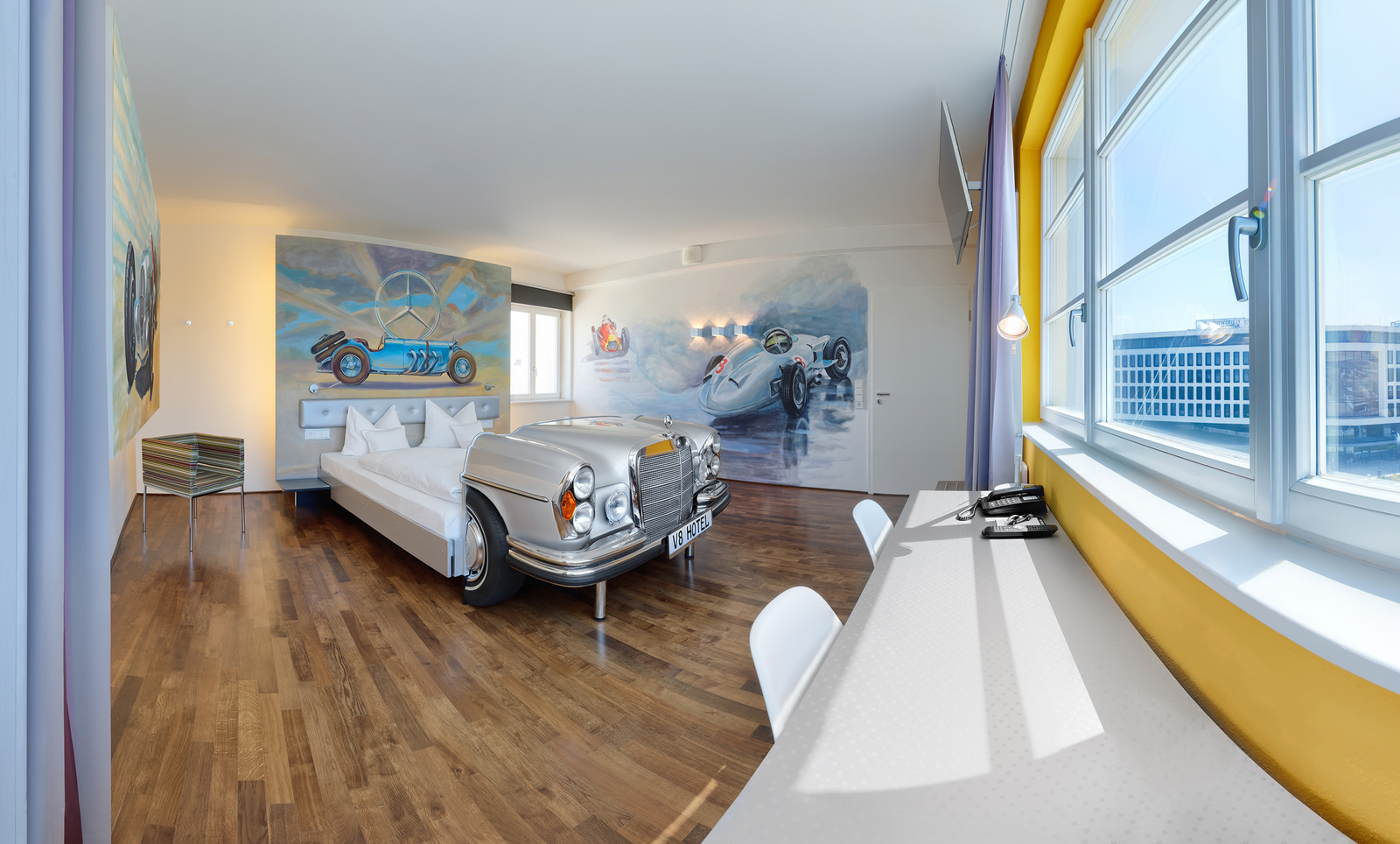 Ein modernes Hotelzimmer mit einem silbernen Auto als Bett. Die Wände sind mit klaren, bunten Autofresken verziert. Es gibt einen Schreibtisch, einen Fernseher und große Fenster, die viel Licht hereinlassen. Der Boden ist aus Holz und die Einrichtung ist minimalistisch und stilvoll.