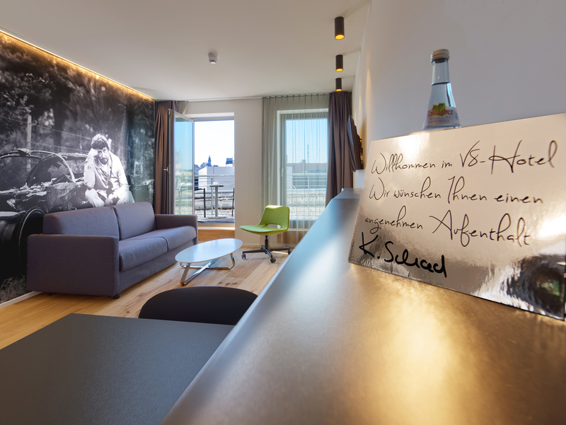 Ein stilvoll eingerichteter Hotelzimmer mit einem grauen Sofa, einem Glastisch und einer modernen grünen Stuhl. An der Wand befindet sich ein großes, monochromes Wandbild. Auf der Arbeitsfläche steht ein Willkommensschild mit der Nachricht, dass ein angenehmer Aufenthalt gewünscht wird.