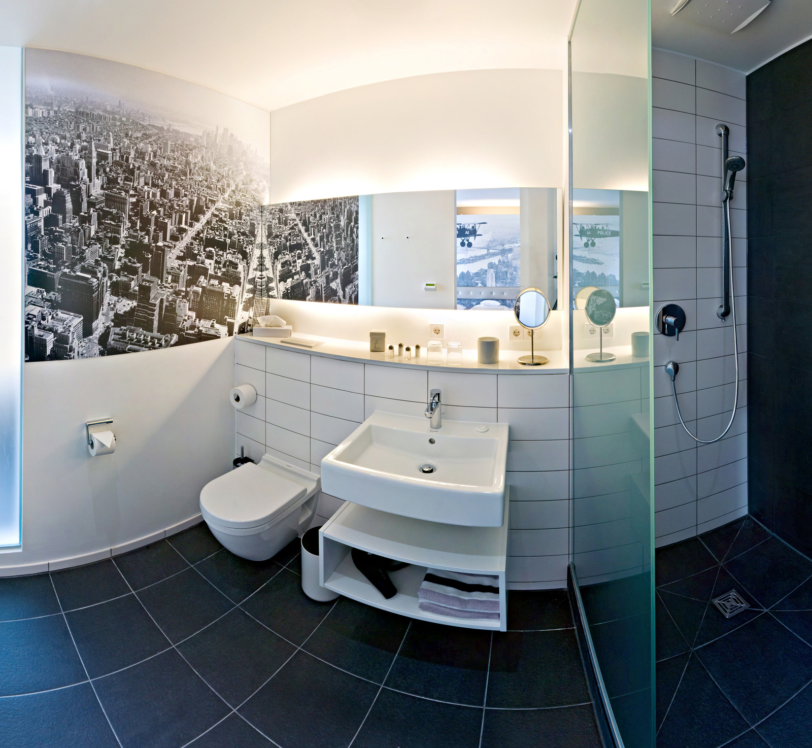 Ein modernes Badezimmer mit einer großen Dusche, einem Waschbecken und einer Toilette. Die Wände sind mit schwarzen und weißen Stadtbildern dekoriert. Eine große Spiegelwand reflektiert Licht und schafft eine helle Atmosphäre. Der Boden ist mit dunklen Fliesen ausgelegt.
