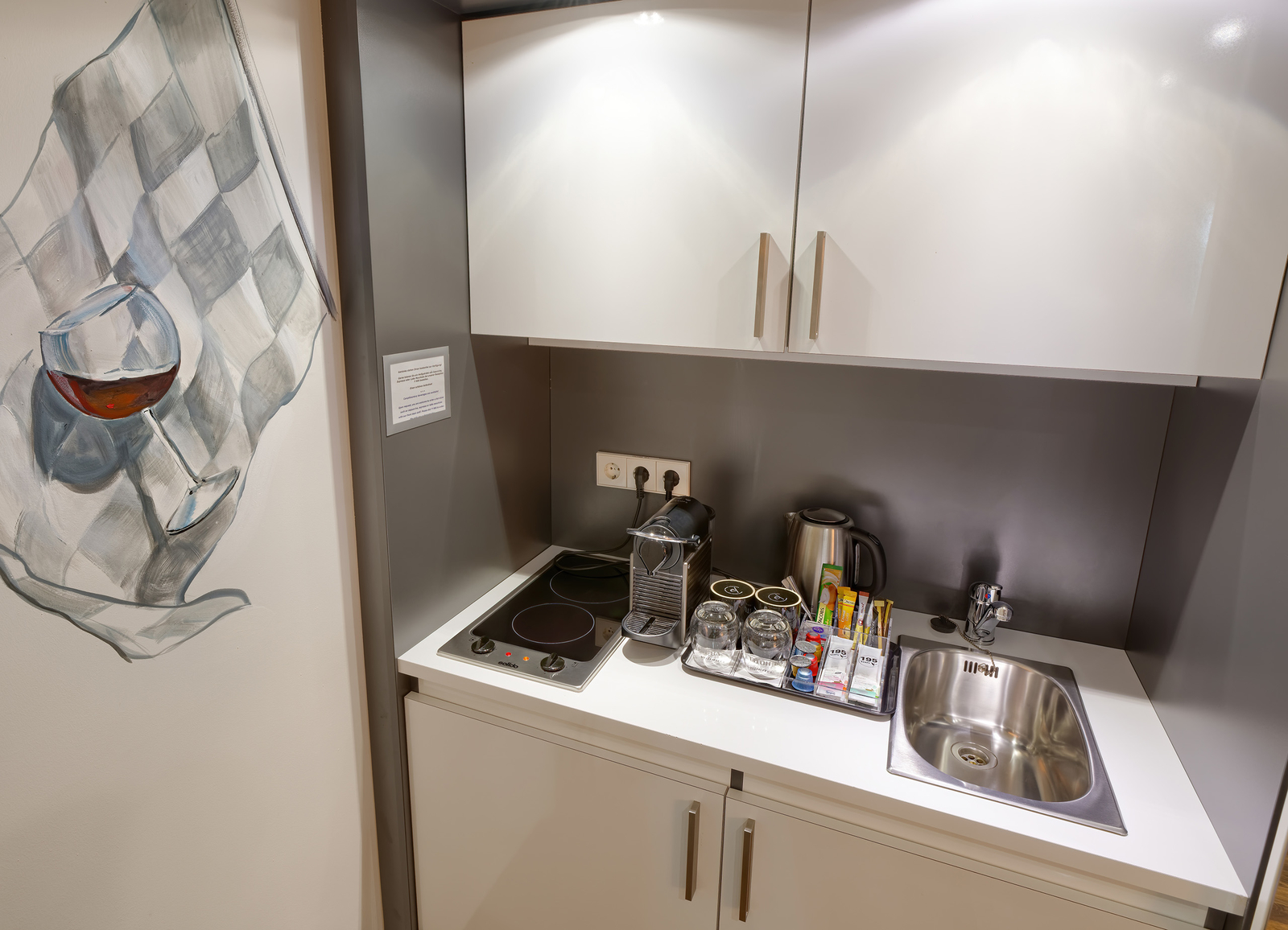 Die Abbildung zeigt eine kleine Küchenzeile mit einem Kochfeld, einer Kaffeemaschine und einem Wasserkocher. Auf der Arbeitsfläche stehen Gläser und mehrere Packungen. An der Wand ist eine stilisierte Darstellung eines Weinglases. Der Raum wirkt modern und funktional.