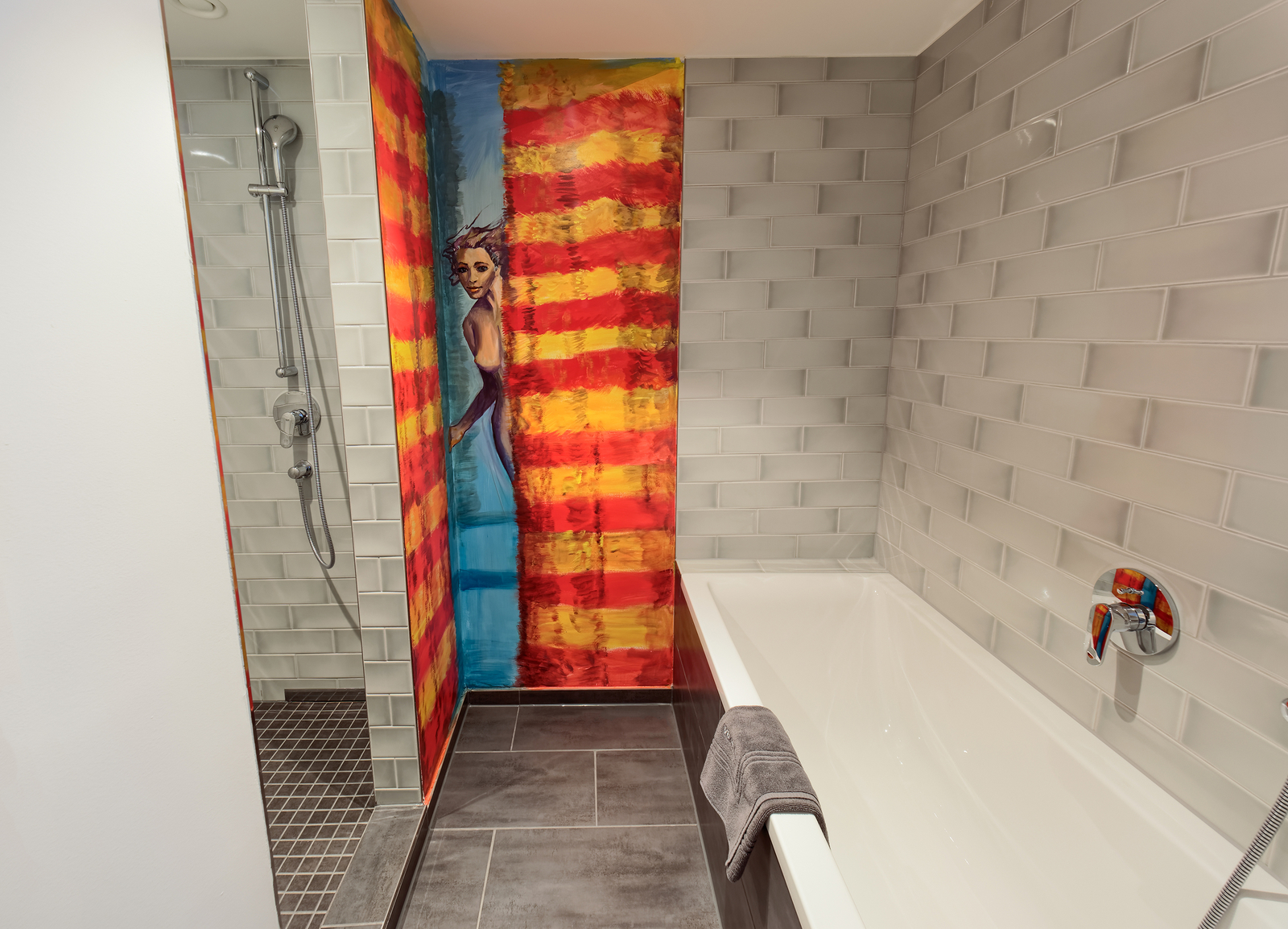 Das Bild zeigt ein Badezimmer mit einer modernen Badewanne und einer Dusche. Eine Wand ist bunt bemalt und zeigt ein künstlerisches Motiv. Die gegenüberliegende Wand ist mit grauen Fliesen gefliest, und ein Handtuch liegt auf der Badewanne.