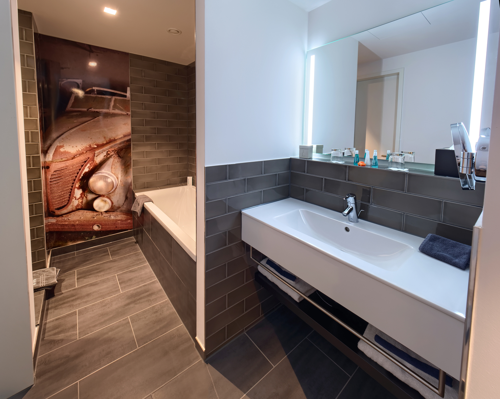 Das Bild zeigt ein modernes Badezimmer mit einer Badewanne und einem Waschbecken. Die Wände sind mit grauen Fliesen verkleidet. Ein großer Spiegel mit beleuchteten Rändern hängt über dem Waschbecken. Im Hintergrund ist ein Wandbild eines alten Autos zu sehen.