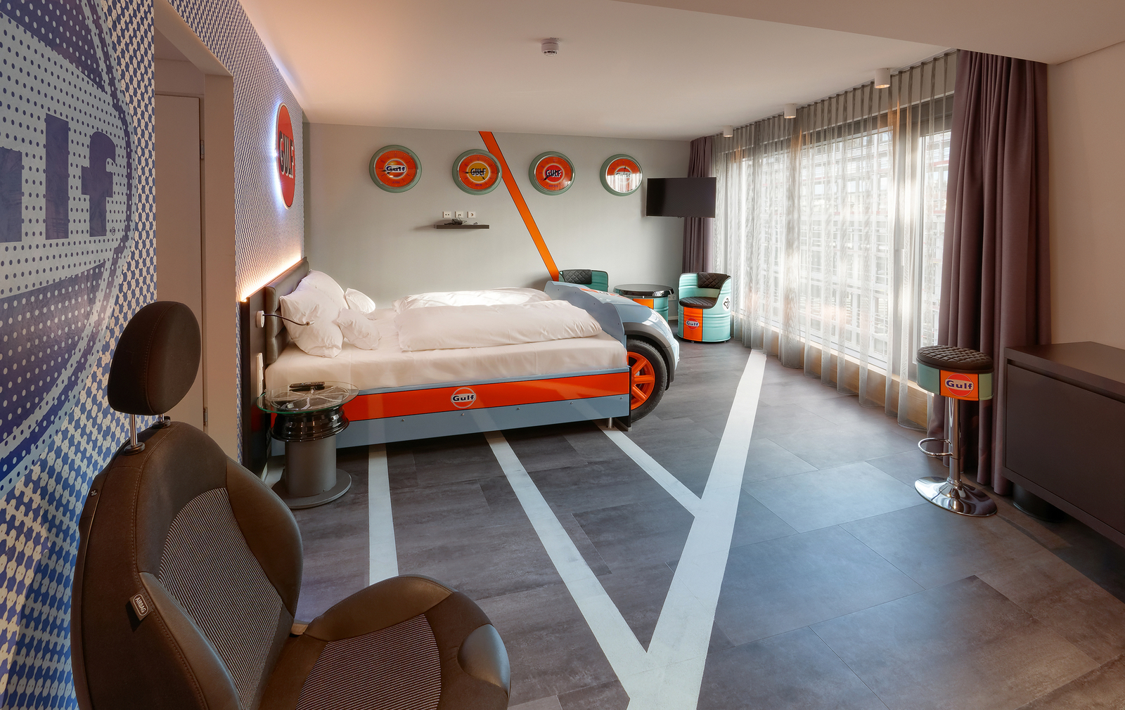 Ein modernes Zimmer mit einem Bett, das in den Farben und dem Design von Gulf gestaltet ist. Die Wände sind mit Gulf-Logos dekoriert. In der Ecke befindet sich ein glasierter Tisch und ein ergonomischer Stuhl. Das Zimmer hat große Fenster mit Vorhängen und bietet eine helle, einladende Atmosphäre.