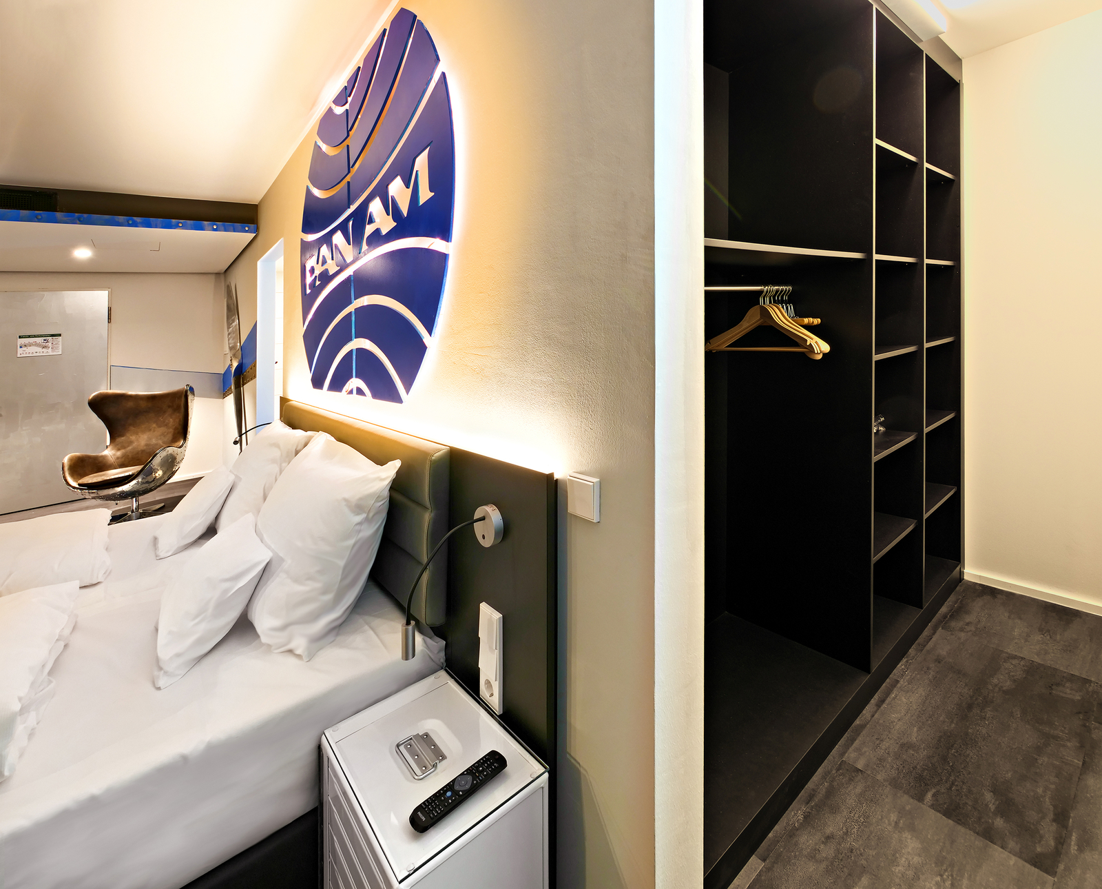 Das Bild zeigt ein modernes Hotelzimmer mit einem Doppelbett, das mit mehreren Kissen ausgestattet ist. An der Wand ist ein leuchtendes Pan Am-Logo zu sehen. Neben dem Bett steht ein kleiner Nachttisch mit einer Lampe und einer Fernbedienung. Im Hintergrund steht ein Sessel und ein offener Kleiderschrank.