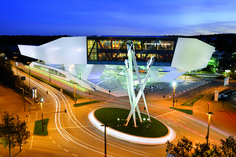 Das Bild zeigt das moderne Porsche Museum in Stuttgart bei Nacht. Die Architektur ist futuristisch, mit einer markanten, geschwungenen Fassade und großen Fenstern. Um das Gebäude verlaufen Straßen, die von Scheinwerferlicht erhellt werden. Impressionen von urbanem Leben unterstreichen die Szene.
