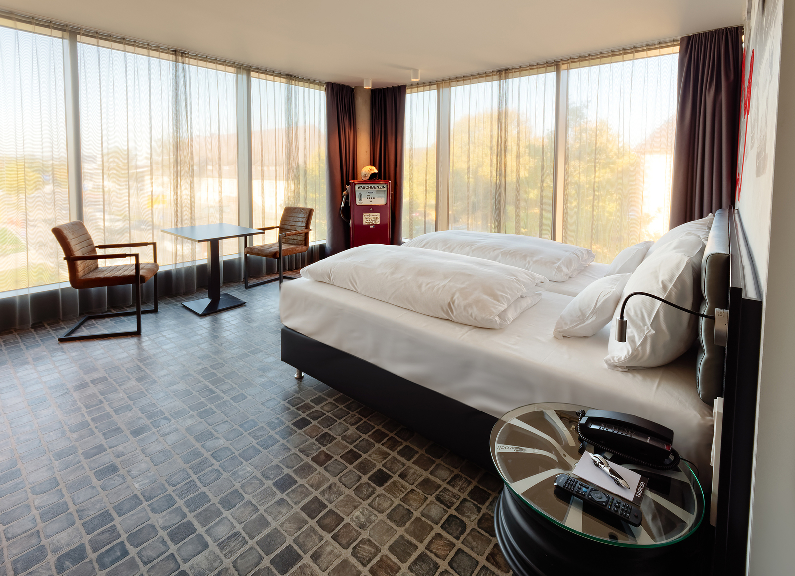 Ein modernes Hotelzimmer mit großen Fenstern, die viel Tageslicht hereinlassen. Im Raum stehen zwei Einzelbetten mit weißen Bettdecken, ein Tisch mit zwei Stühlen sowie ein Nachttisch mit einem Telefon und einer Fernbedienung. Der Boden ist mit grauen Pflastersteinen ausgelegt.