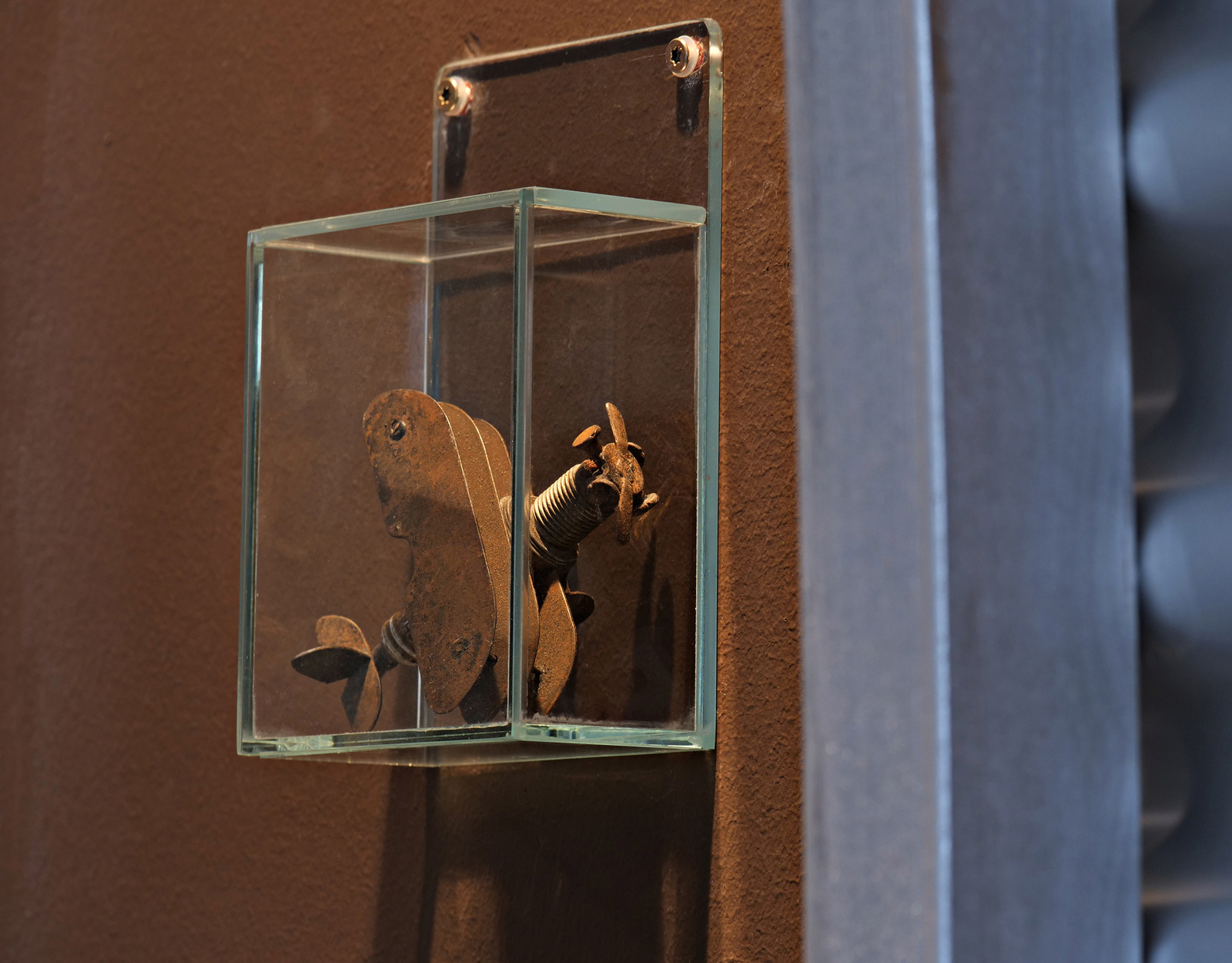 Das Bild zeigt eine Glasvitrine, in der sich ein altes, rostiges mechanisches Teil befindet. Es hat die Form eines Propellers und einige bewegliche Komponenten, die künstlerisch angeordnet sind. Der Hintergrund ist in einem neutralen Braunton gehalten.
