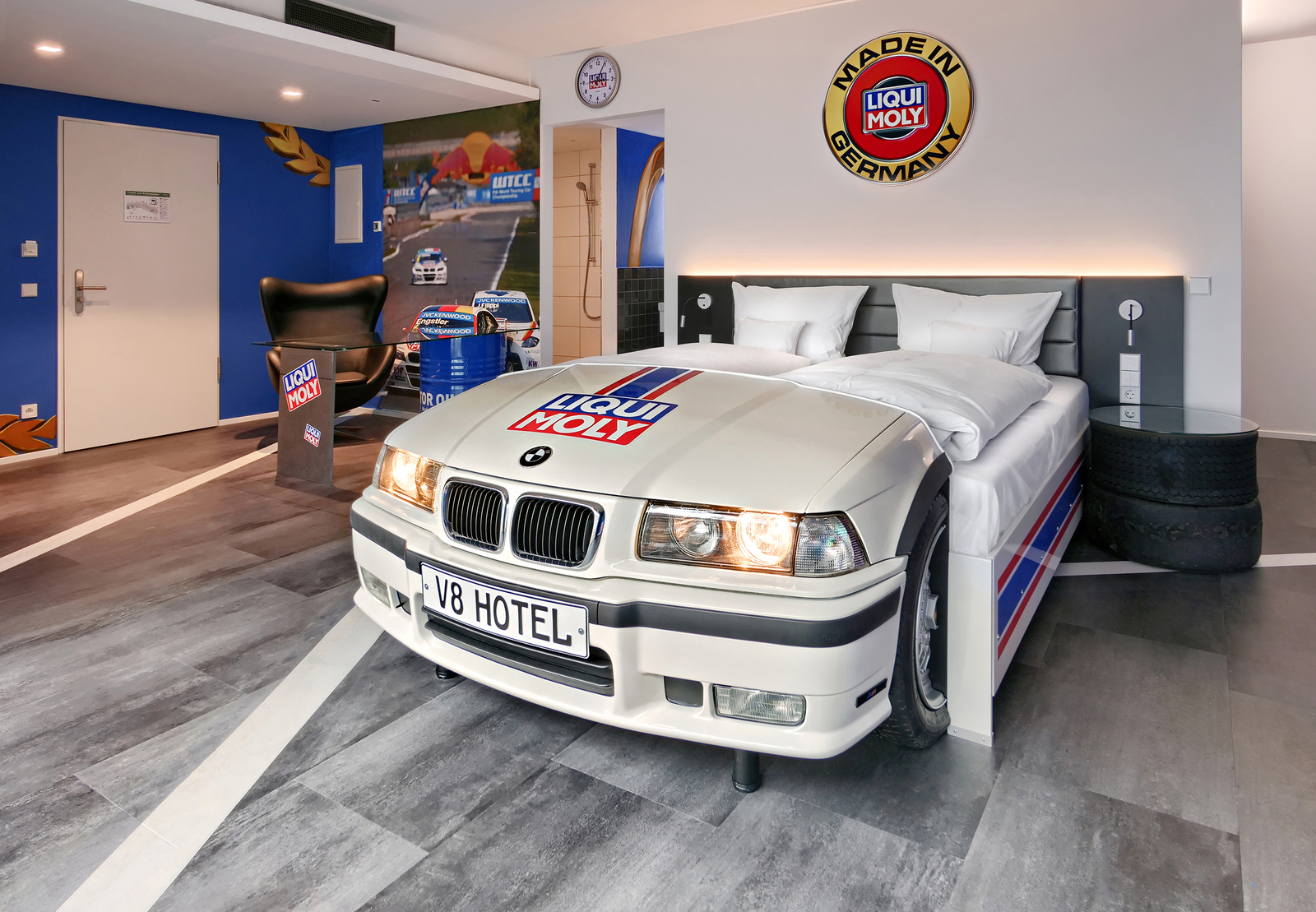 Das Bild zeigt ein Hotelzimmer mit einem Bett, das die Vorderseite eines weißen BMW-Rennwagens imitiert. Der Wagen ist mit einem Liqui Moly-Logo versehen. Im Hintergrund sind weitere Elemente eines Rennsport-Themenzimmer zu sehen, einschließlich eines Monitors und dekorativer Grafiken.