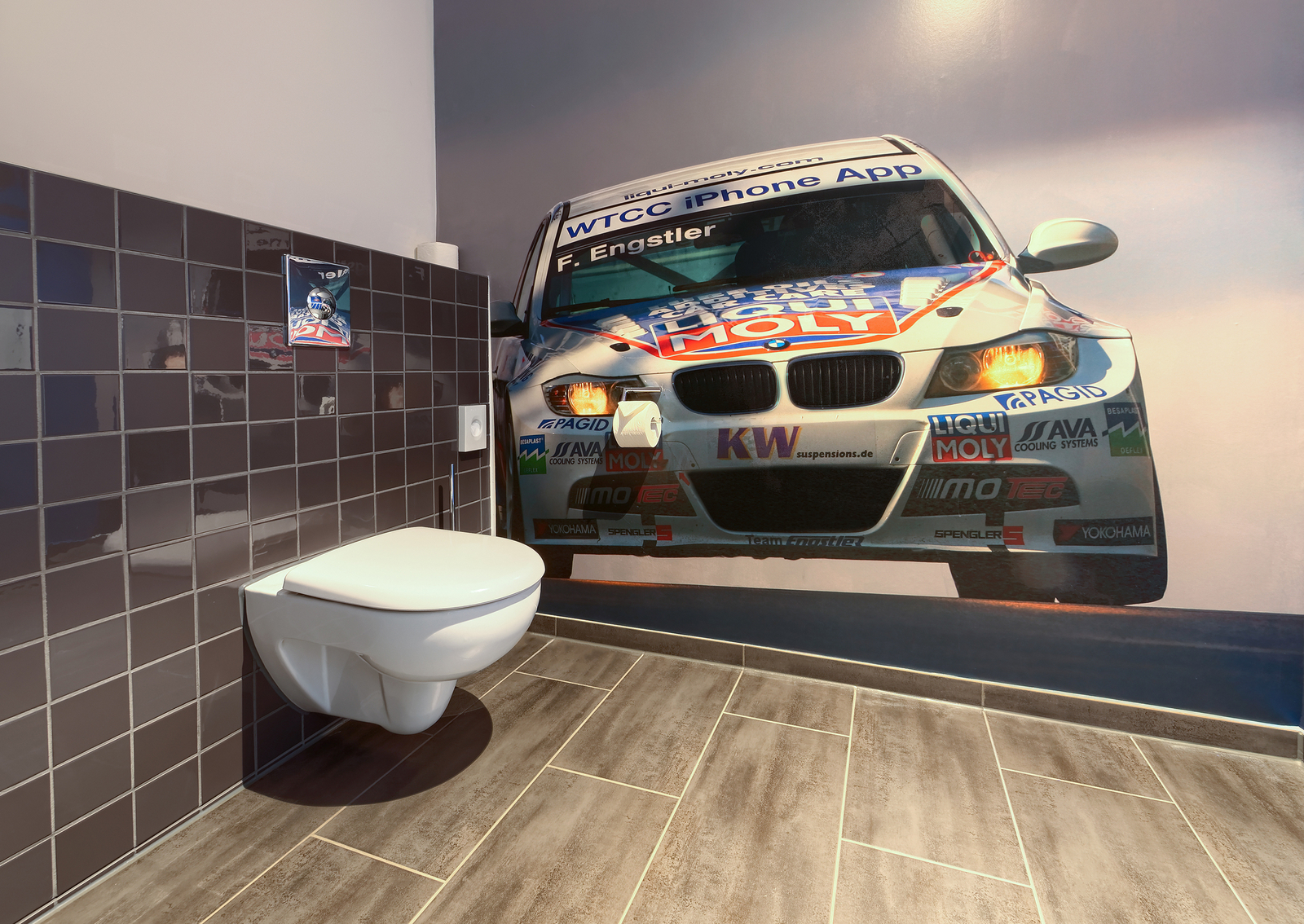 In einem modernen Badezimmer steht eine weiße Wandtoilette vor einer großflächigen Fotowand, die ein Rennfahrzeug zeigt. Die Fußbodenfliesen sind in einem ansprechenden Holzlook gestaltet, und die Wände sind mit dunklen Fliesen versehen.