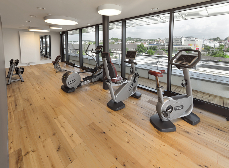 Heller Fitnessraum mit bodentiefen Fenstern und mehreren Sportgeräten davor.