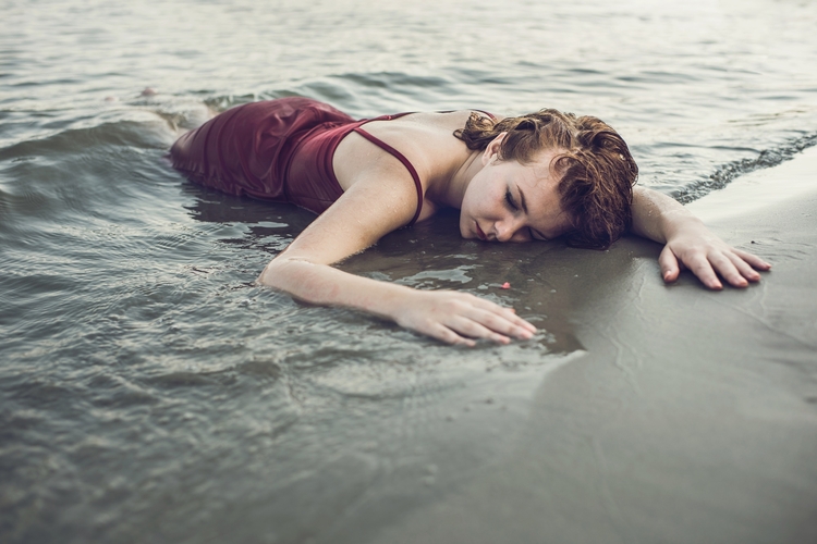 Eine Person liegt auf dem Sand, während Wasser über sie fließt. Sie hat braune Haare und trägt ein rotes Kleid. Der Gesichtsausdruck deutet auf Ruhe oder Nachdenklichkeit hin. Im Hintergrund ist eine ruhige Wasseroberfläche zu sehen, die die Stimmung verstärkt.