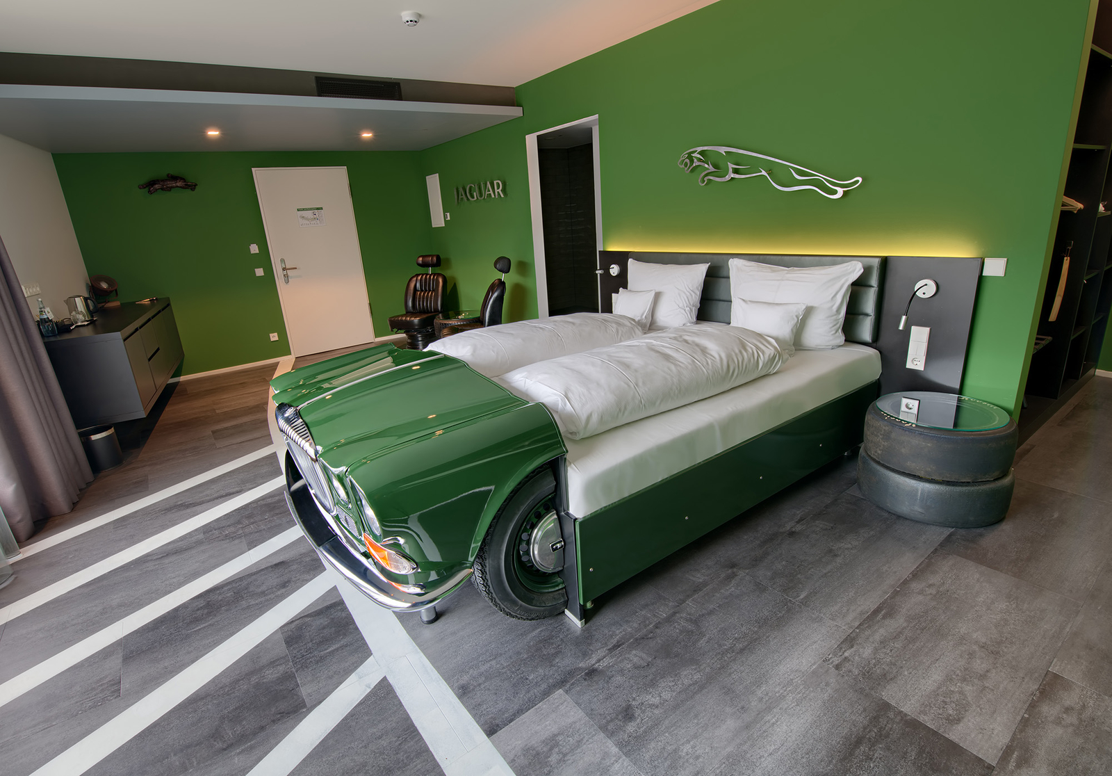 Das Bild zeigt ein kreatives Zimmerdesign mit einer grünen Wand. Im Mittelpunkt steht ein Bett, das wie die Vorderseite eines Autos gestaltet ist, mit einer eleganten Motorhaube und Rädern. Es gibt zusätzliche Möbel wie einen Sessel und einen Tisch aus Autoreifen.