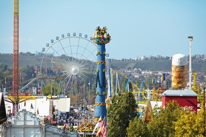 Eine belebte Jahrmarkt-Szene mit Fahrgeschäften im Hintergrund. Im Vordergrund steht ein geschmückter, hoher Turm. Links ist die Fliegende Riesenschaukel zu sehen, während ein großes Riesenrad im Hintergrund dreht. Die Atmosphäre wirkt festlich und einladend.
