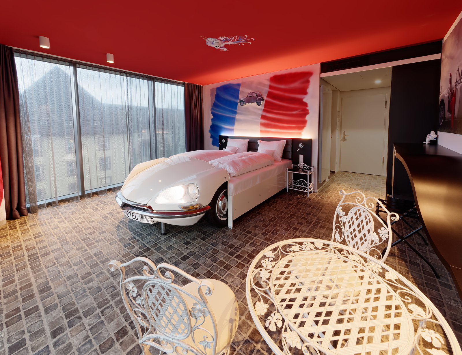 Das Bild zeigt ein kreatives Hotelzimmer mit einem Bett in Form eines weißen Autos, das an der Wand eine künstlerische Darstellung in den Farben der französischen Flagge hat. Der Raum hat große Fenster und ist stilvoll mit einem eleganten Tisch und Stühlen eingerichtet.