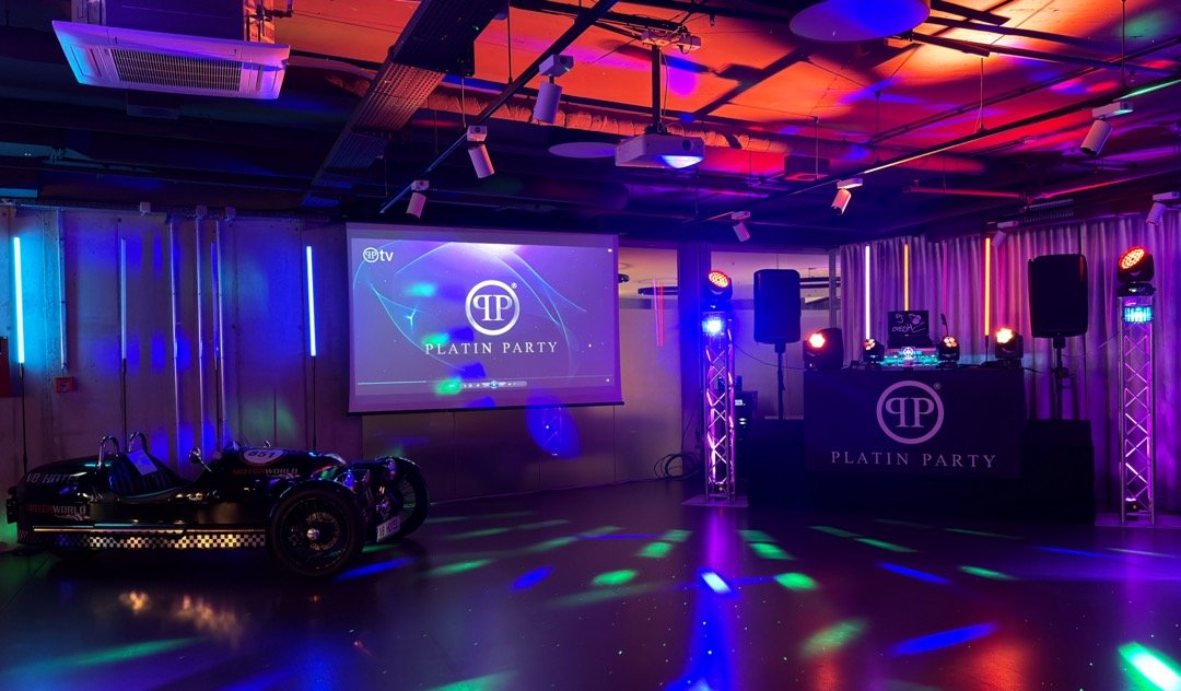 Ein Veranstaltungsraum mit einem DJ-Pult und einer Projektorleinwand, auf der "PLATIN PARTY" steht. Links steht ein schwarz-weißes Auto. Der Raum ist mit bunten Lichtern beleuchtet, die eine festliche Atmosphäre schaffen.