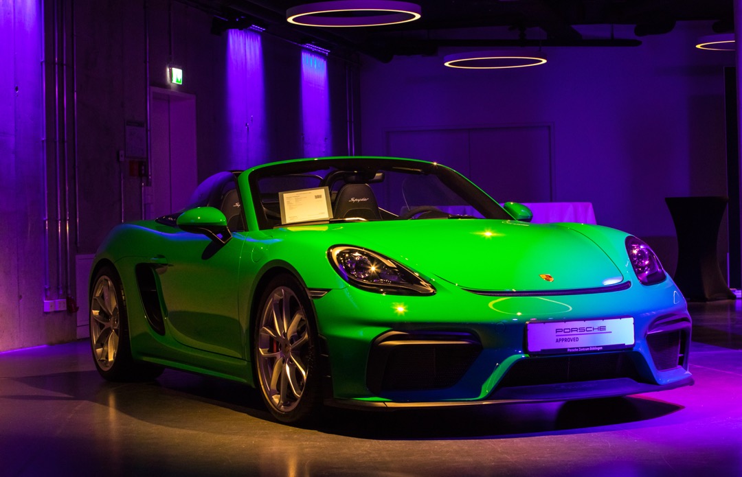 Ein leuchtend grüner Sportwagen, ein Porsche 718 Spyder, steht in einem modern gestalteten Raum mit sanfter Beleuchtung. Der Wagen hat ein offenes Dach und auffällige Felgen. Ein Informationsblatt ist auf dem Armaturenbrett platziert, das Details zum Fahrzeug enthält.