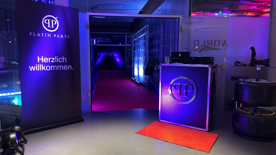 Ein Empfangsbereich bei einer Veranstaltung. Links steht ein Banner mit der Aufschrift „Platin Party – Herzlich willkommen“. Im Hintergrund führt ein roter Teppich zum V8 Hotel, das mit blauen und violetten Lichtern beleuchtet ist. Rechts befindet sich ein Empfangstisch.