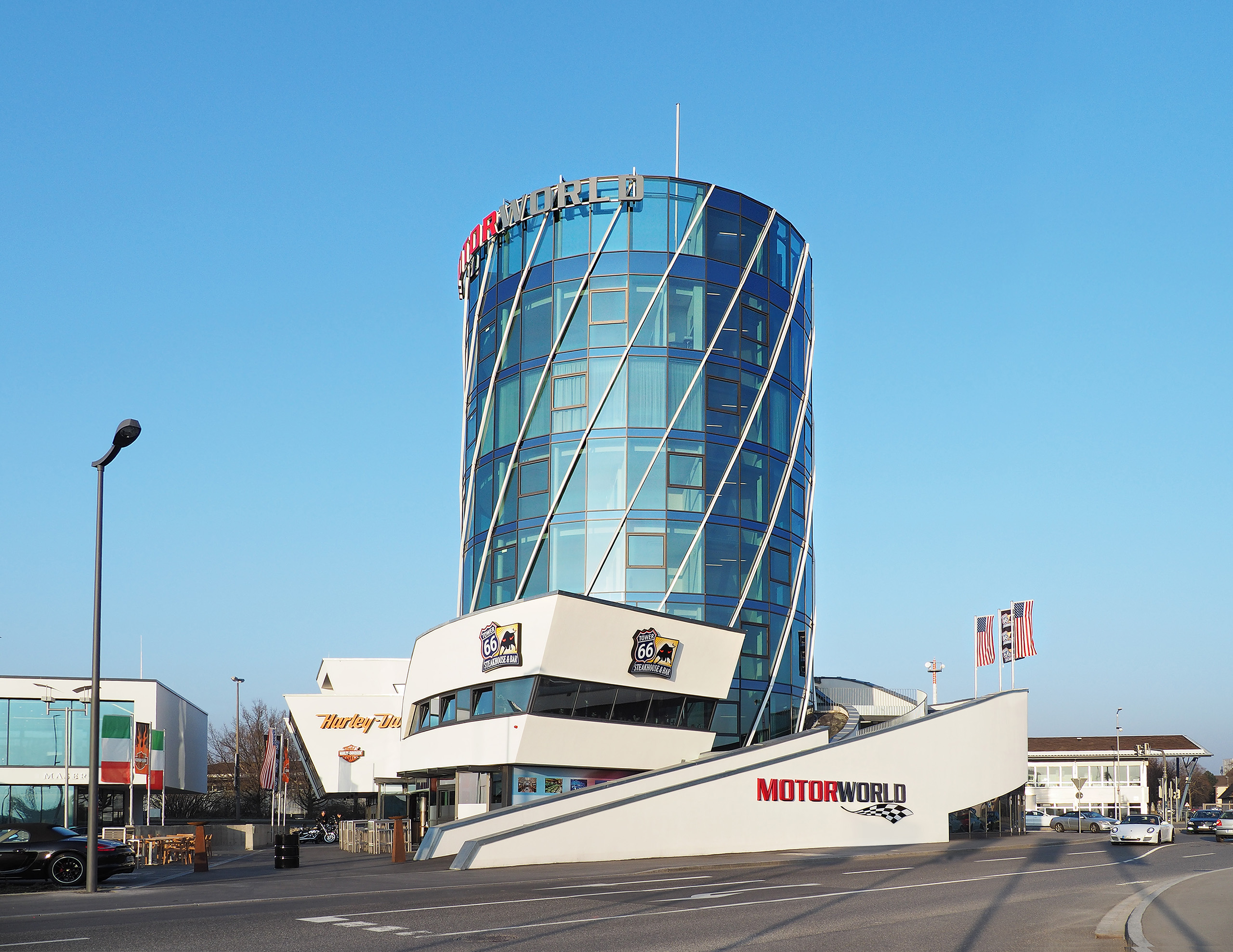 Das Bild zeigt ein modernes, gläsernes Gebäude mit einer einzigartigen, schrägen Fassade. Vor dem Gebäude befindet sich eine breite Treppe mit dem Schriftzug MOTORWORLD. Im Hintergrund sind weitere Bauwerke sichtbar, darunter Restaurants und Flaggen. Der Himmel ist klar und blau.