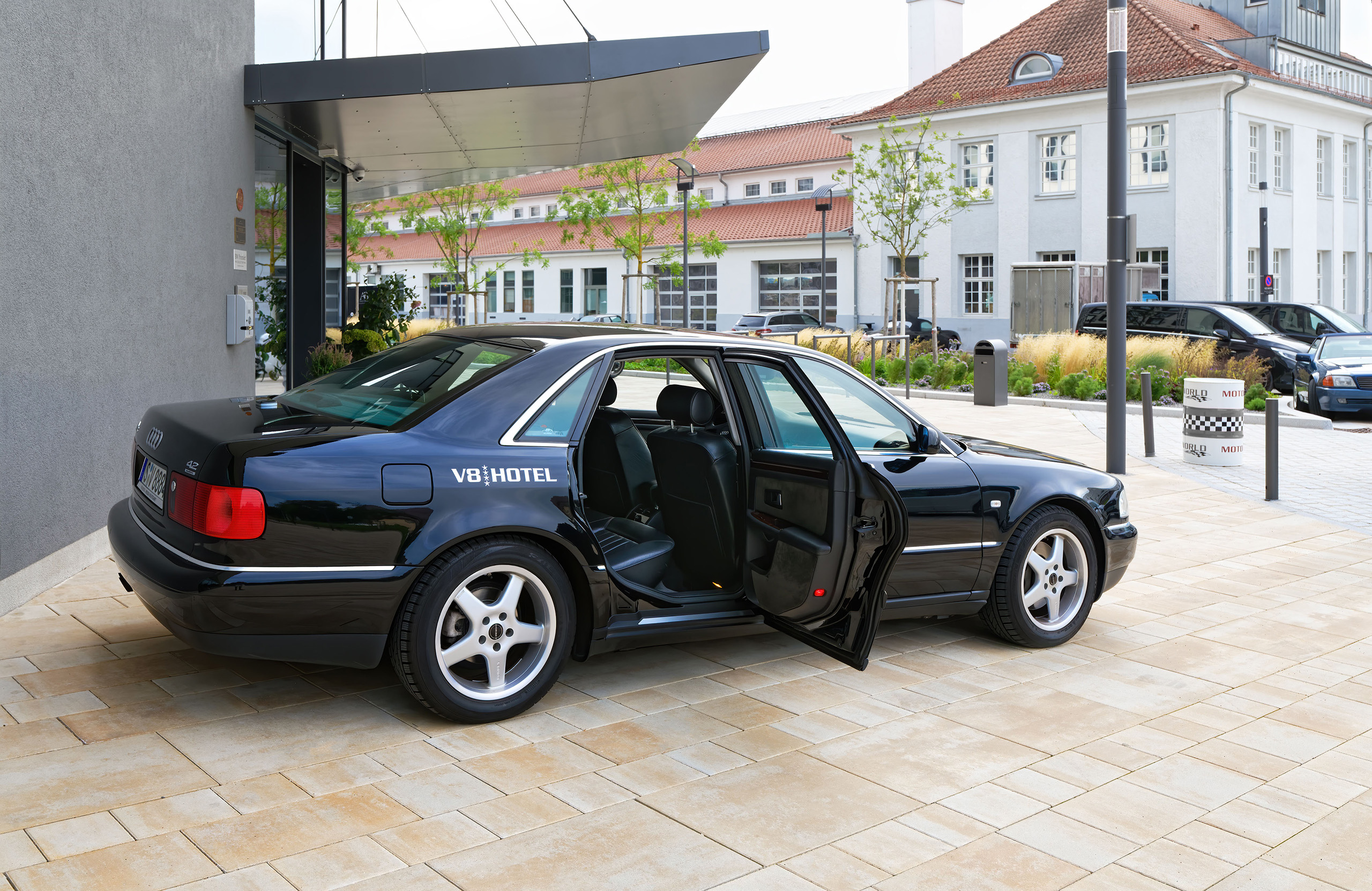 Ein schwarzes Auto mit offenem Türen steht vor einem modernen Gebäude. An der Seite des Fahrzeugs ist der Schriftzug V8 HOTEL zu sehen. Der Parkplatz ist gepflastert, mit einigen Pflanzen im Hintergrund und weiteren Autos in der Umgebung.
