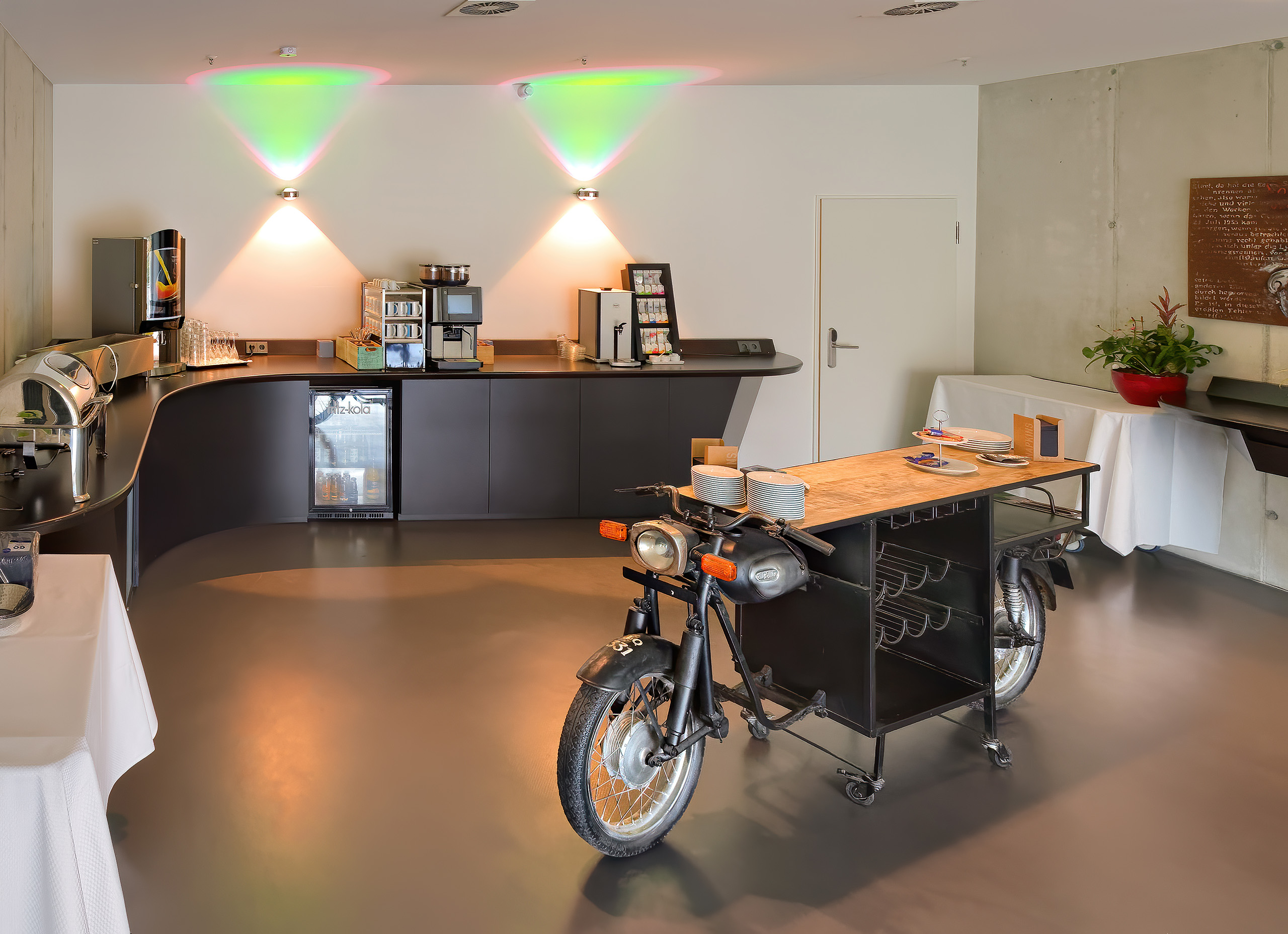 Im Bild ist eine moderne Café-Bar zu sehen. Links befindet sich ein geschwungener Tresen mit verschiedenen Getränken und Snacks. In der Mitte steht ein Tisch, der aus einer umgebauten Oldtimer-Motorradplatte besteht. Die Beleuchtung ist farbenfroh und modern gestaltet.
