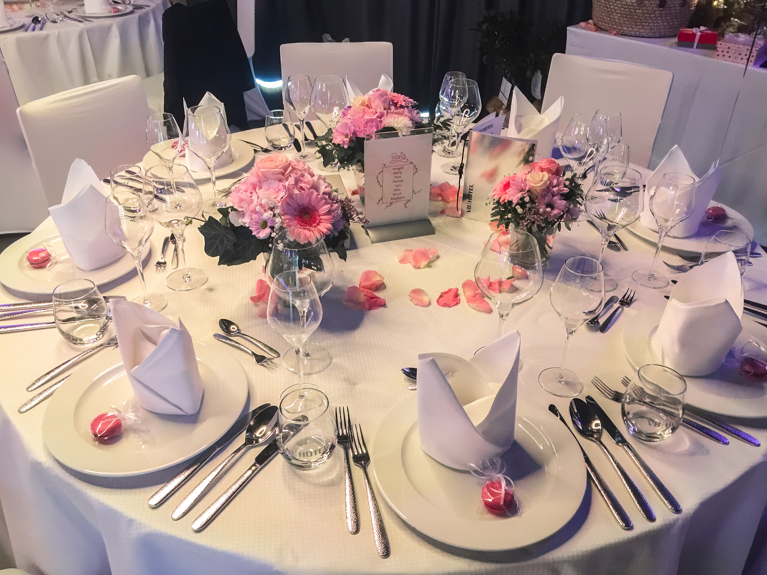 Ein eleganter festlich gedeckter Tisch mit weißen Tischdecken. Es sind Teller, Gläser und Besteck perfekt angerichtet. Mittig stehen kreative Blumenarrangements in Rosa und Weiß, umgeben von Blütenblättern. Kleine Bonbons liegen auf den Tellern, die insgesamt einladend und dekorativ wirken.
