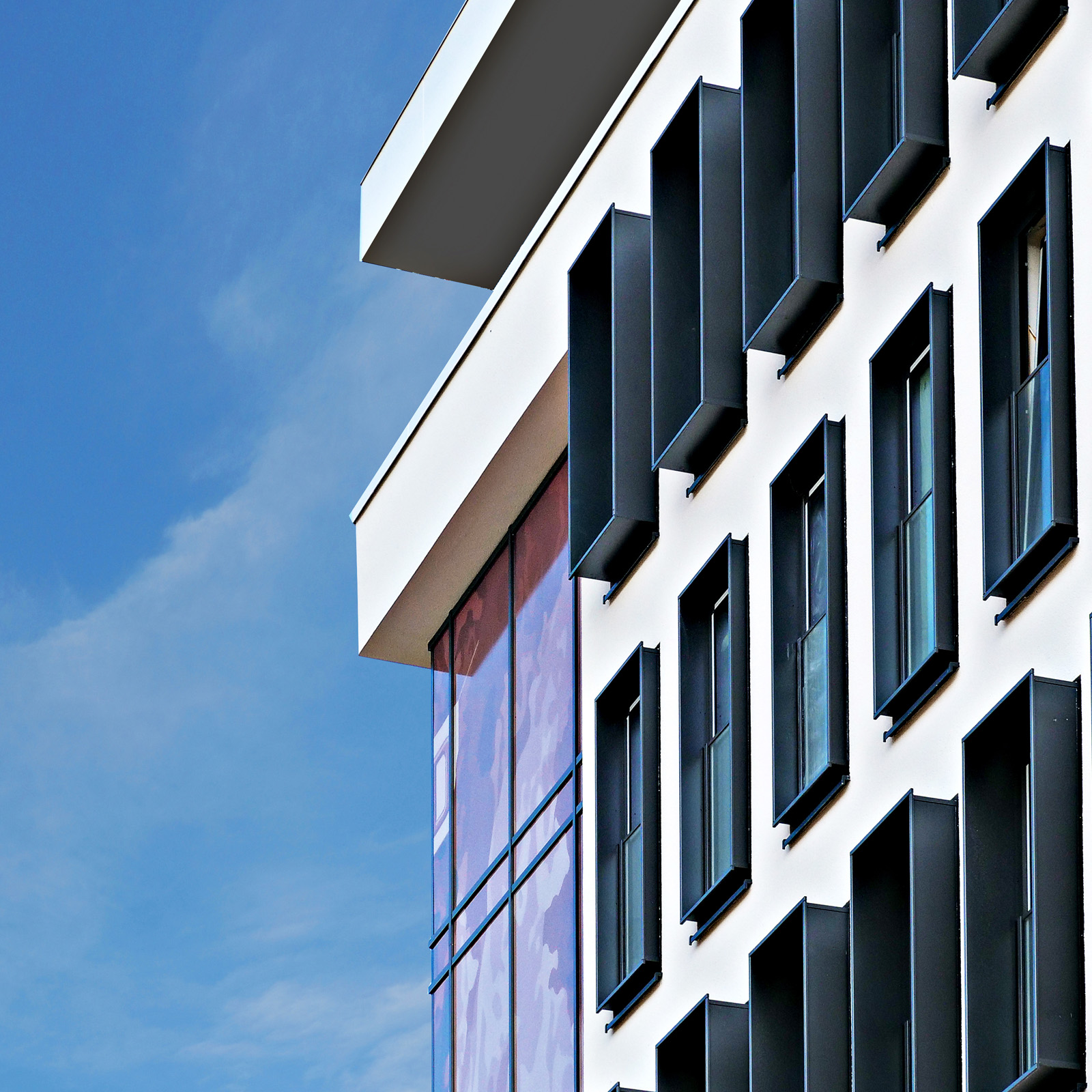 Ein moderner Gebäudewinkel mit weißen Wänden und schwarzen Fensterläden. Die Fassade zeigt eine Kombination aus Fenstern und einer gläsernen Fläche, die farblich hervorsticht. Im Hintergrund ist der blaue Himmel sichtbar.