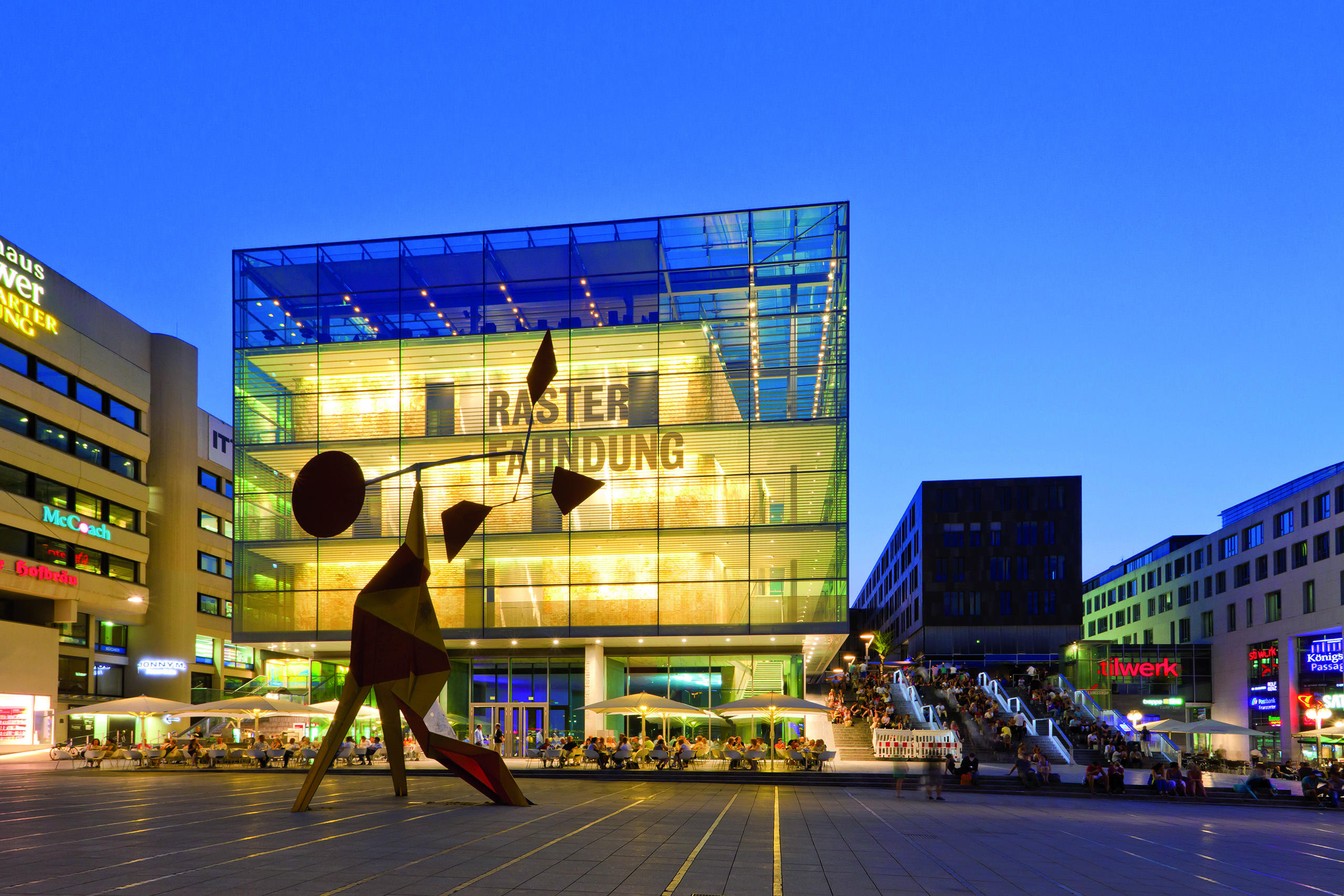 Das Stuttgarter Kunstmuseum. Davor steht eine abstrakte, farbenfrohe Skulptur. Um das Gebäude sind Tische und Stühle auf einem Platz angeordnet, und im Hintergrund sind Menschen an Treppen und in Geschäften zu sehen. Es ist eine blaue Abenddämmerung.