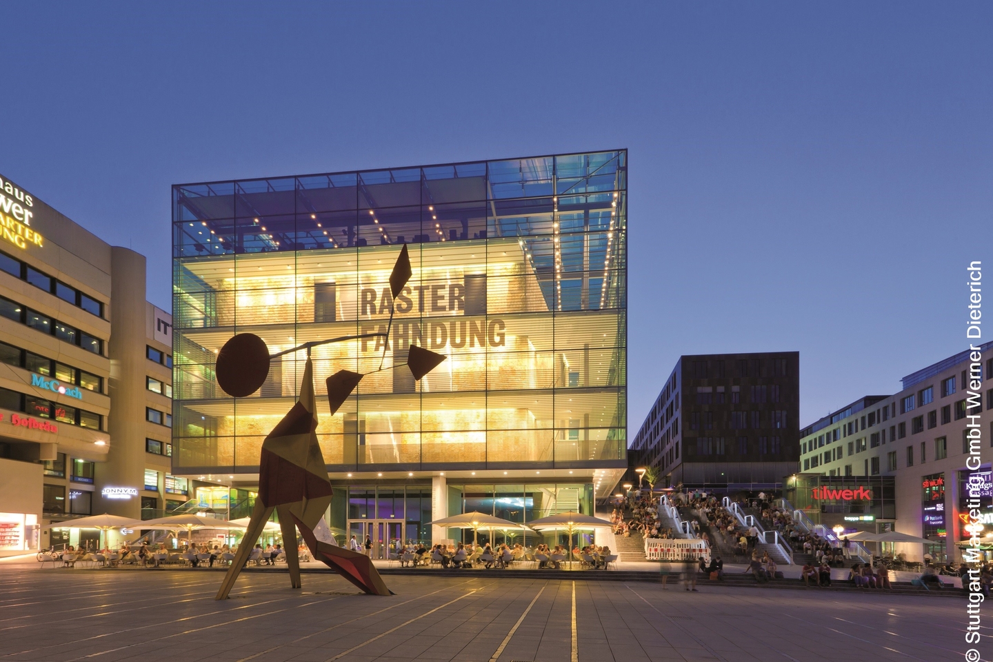 Das Glasgebäude vom Kunstmuseum Stuttgart mit seinem großen Vorplatz, auf dem eine moderne Skulptur steht.