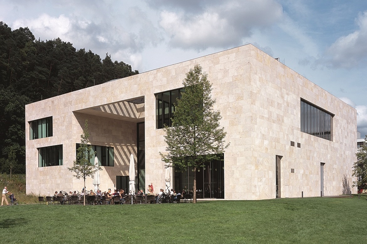 Ein modernes zweigeschossiges Gebäude aus hellem Stein, umgeben von Wiese. Die Fassade hat große Fenster. Vor dem Gebäude sitzen mehrere Personen an Tischen, viele von ihnen genießen im Freien. Im Hintergrund sind Bäume und ein bewölkter Himmel sichtbar.