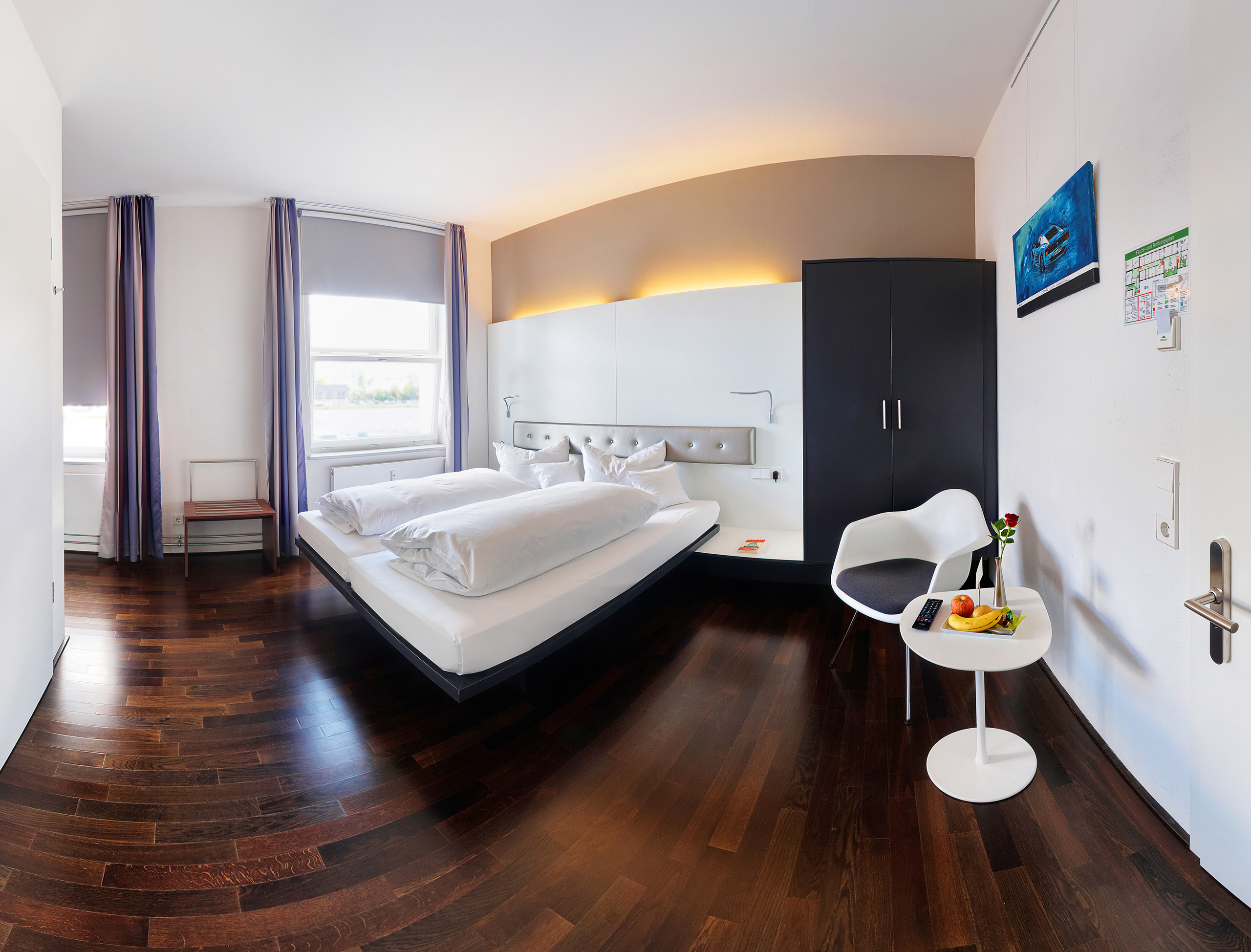Ein modernes, barrierefreies Hotelzimmer mit einem Doppelbett, zwei Kissen und einem Nachttisch. Neben dem Bett steht ein kleiner Tisch mit einer Blume, einem Obstkorb und einer Fernbedienung. Große Fenster lassen Tageslicht herein, und der Boden ist aus Holz.
