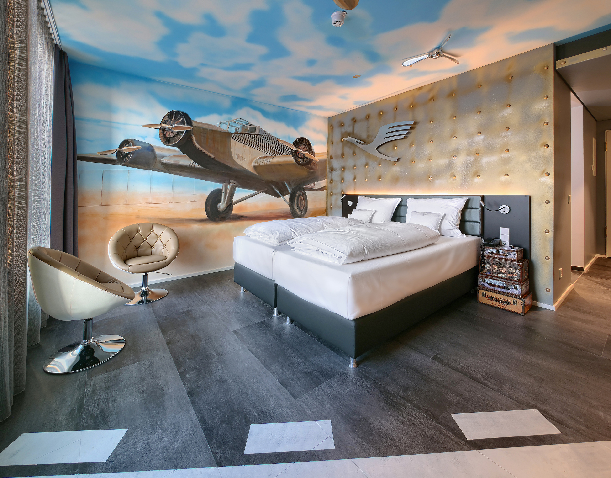 Gemütliches Doppelbett und zwei runde Sessel im Airfield-Themenzimmer im V8 Hotel mit einer Motivtapete, die ein Propellerflugzeug zeigt