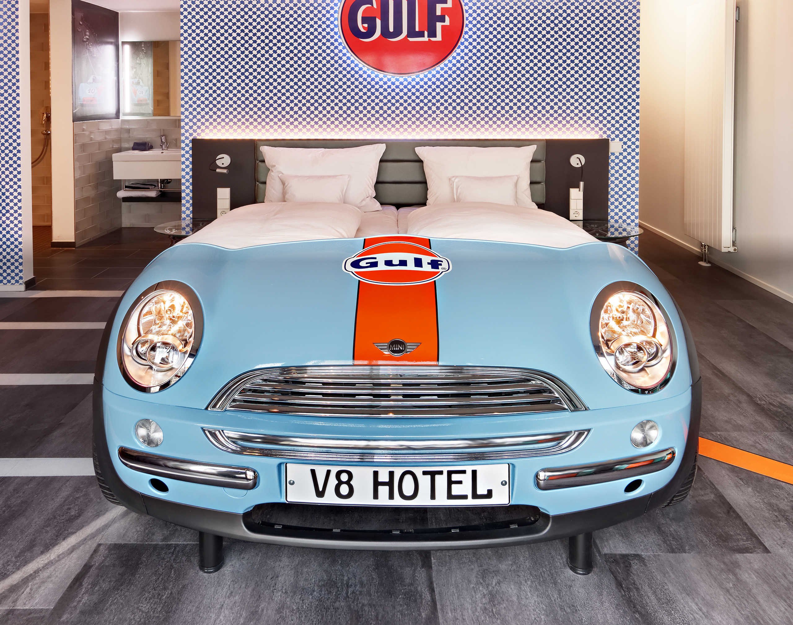 Blick auf ein hellblaues Autobett der Marke Mini an einer karierten Wand mit dem orangefarbenen Gulf-Logo