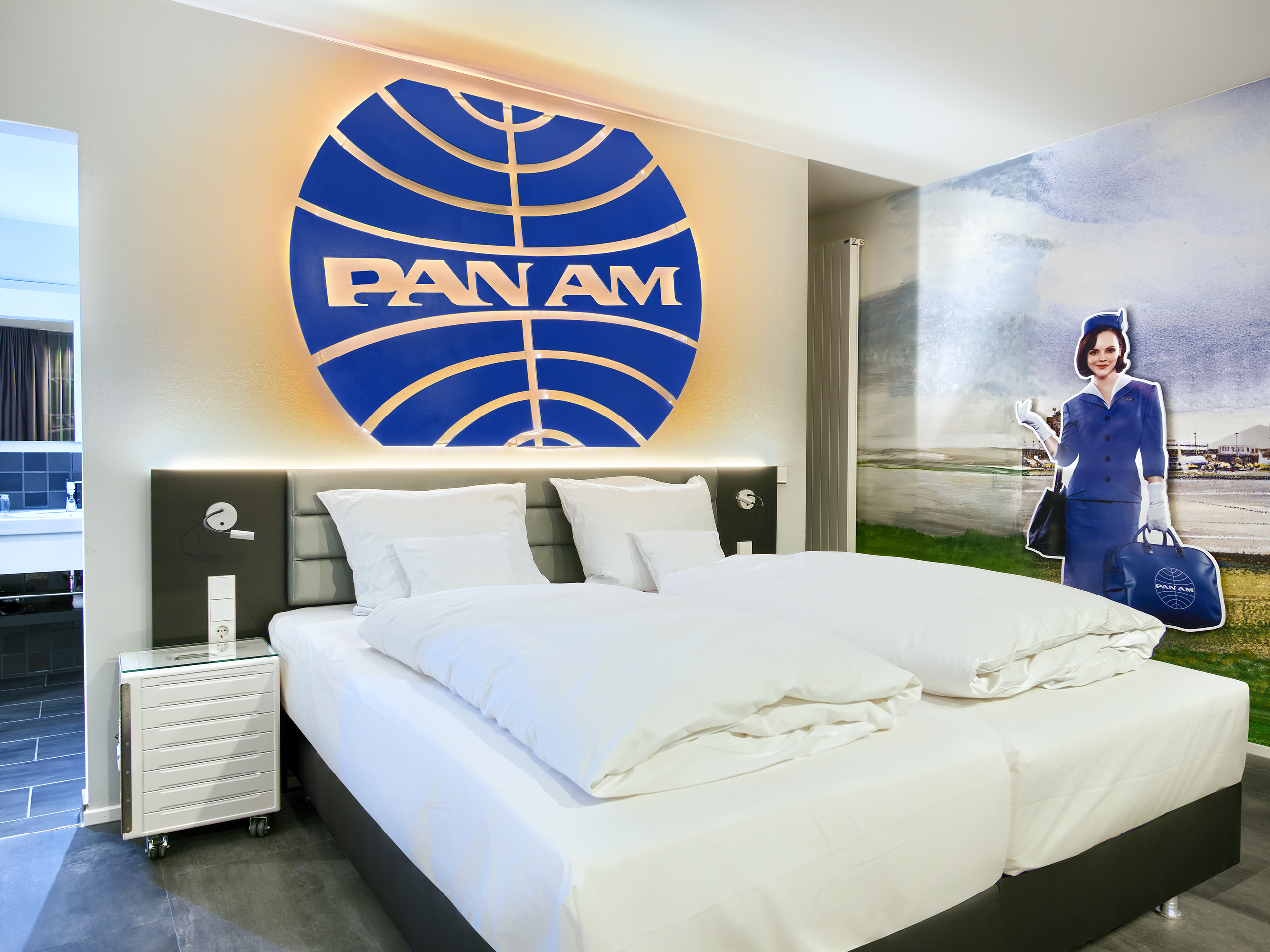 Das Bild zeigt ein modernes Hotelzimmer mit einem großen PAN AM-Logo an der Wand. Im Hintergrund steht eine Figur einer Stewardess in einem blauen Uniformanzug mit einer Tasche. Das Zimmer ist hell gestaltet, mit einem großen Doppelbett und eleganten Möbeln.
