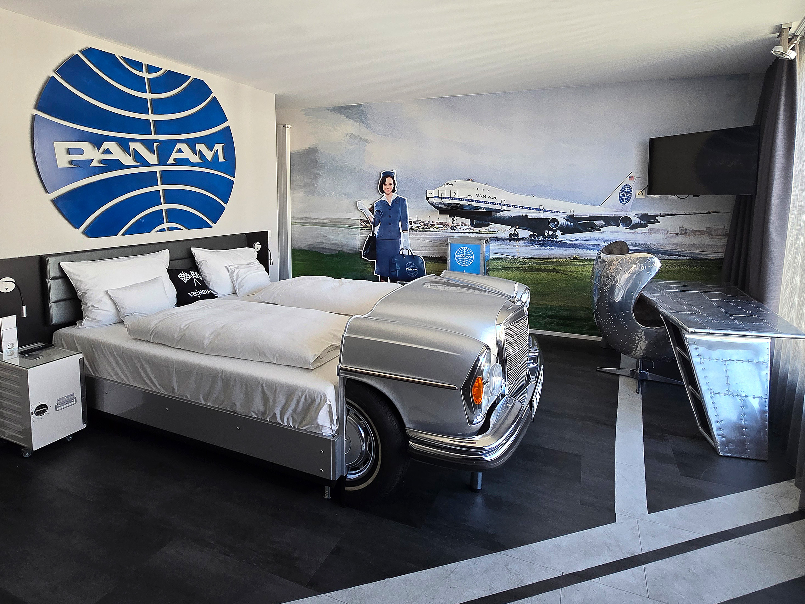 Das Bild zeigt ein Pan Am-Themenzimmer mit einem Bett in Form eines Autos. An der Wand ist ein großes Pan Am-Logo sowie ein Bild eines Flugzeugs. Es gibt auch einen schicken Schreibtisch und eine Figur einer Flugbegleiterin. Das Design ist nostalgisch und erinnert an die Luftfahrtgeschichte.