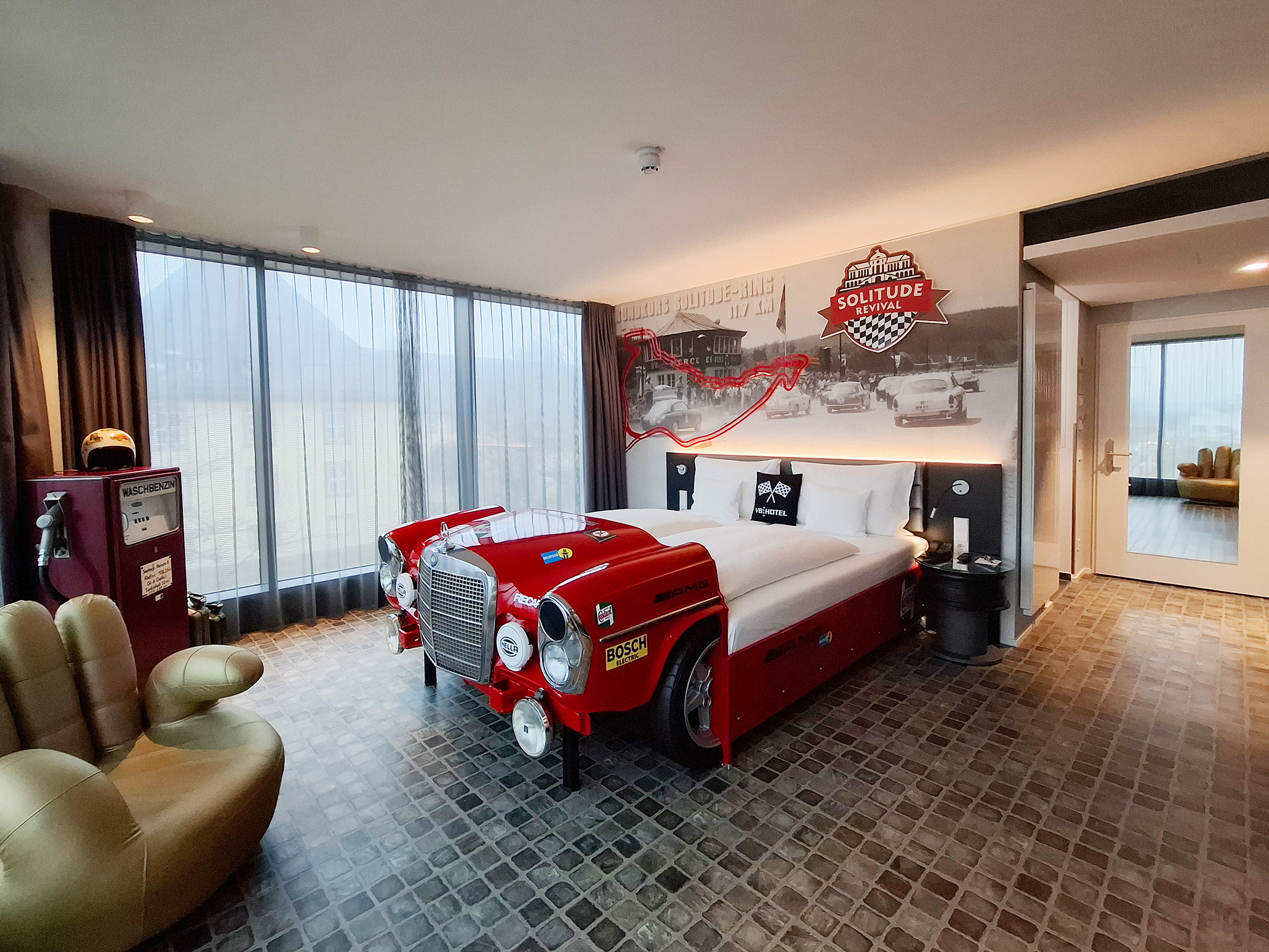 Das Bild zeigt ein stylisches Hotelzimmer mit einem Bett, das die Form eines roten Rennwagens hat. An der Wand ist ein nostalgisches Auto-Motiv. Zudem ist ein bequemer Sessel in Form einer Hand zu sehen. Das Zimmer vermittelt ein modernes, sportliches Ambiente.
